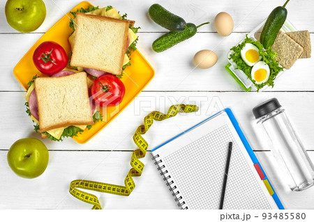 Top view planning notebook with copy space and healthy breakfast. Sandwich with ham and cheese, eggs, cucumber, tomato and bottle of water on white wooden table. 93485580