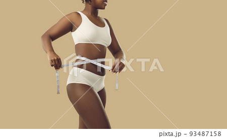 Slender woman who loses weight measures her waist with measuring tape standing on beige background. 93487158