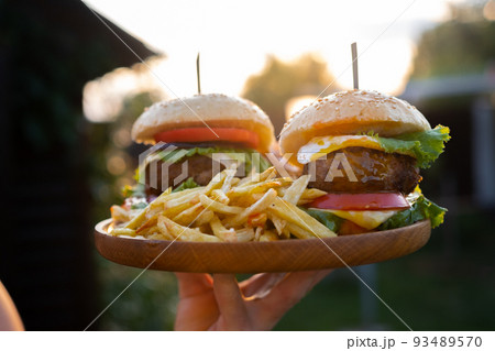 Two hamburgers with beef burger cutlet, fried onion, ketchup sauce and cheese in traditional buns, served on wood chopping board. A hand holds a tray in the open air of the sunset 93489570