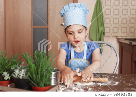 5 year old girl in blue dress and dressed as chef in kitchen laughs and rolling out dough 5 year old girl in blue dress and dressed as chef in kitchen laughs and rolling out dough 93493202