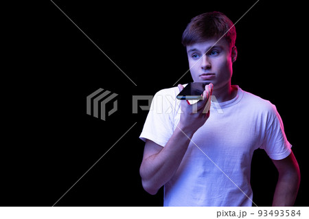 Portrait of young man in white T-shirt posing, recording voice message on phone isolated over black background in neon light Portrait of young man in white T-shirt posing, recording voice message on phone isolated over black background in neon light 93493584