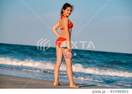 Sandy tanned woman with snorkeling equipment on the beach background. Happy lifestyle. Joyful woman in bikini standing along the beach to the sea. Sandy tanned woman with snorkeling equipment on the beach background. Happy lifestyle. Joyful woman in bikini standing along the beach to the sea. 93496803