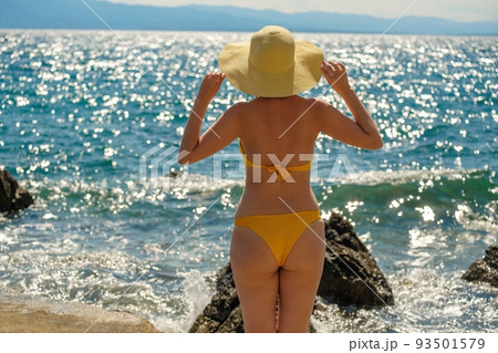 Woman in yellow bikini and straw hat looks at blue sea on warm day. Lady holds headdress with hands against water sparkling in sun close back view 93501579