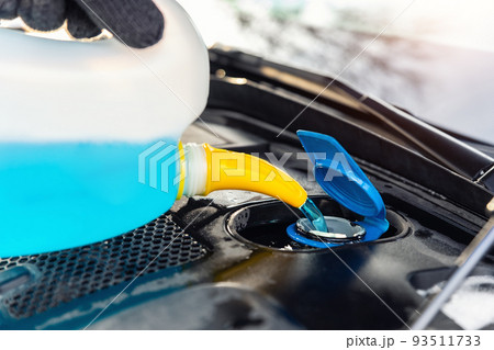 Close-up detail of driver or mechanic hand in gloves pouring blue antifreeze liquid for winter car windshield screen washing with bootle watering can. Car seasonal service and maintenance concept 93511733