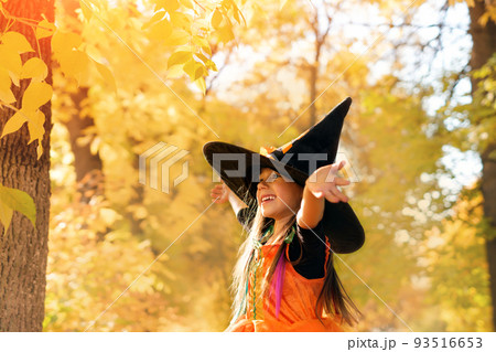 Trick or treating. Portrait of a laughing, cheerful girl in a witch costume and a big black hat on a background of yellow leaves. The child looks away with a smile and an open mouth. halloween Trick or treating. Portrait of a laughing, cheerful girl in a witch costume and a big black hat on a background of yellow leaves. The child looks away with a smile and an open mouth. halloween 93516653