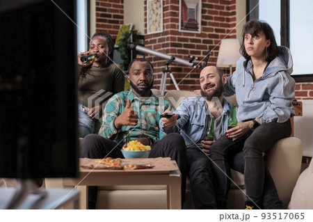 Cheerful people watching tv show or movie on television, gathering together at home. Drinking beer bottles and eating snacks, looking at film on tv channel program, entertainment. 93517065