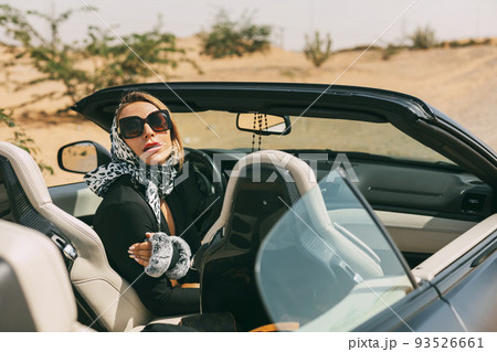 Glamorous blonde in a silk scarf and sunglasses in the driver's seat of a convertible convertible on a hot desert background. Fashion, style, glamour Glamorous blonde in a silk scarf and sunglasses in the driver's seat of a convertible convertible on a hot desert background. Fashion, style, glamour 93526661