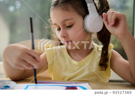 Close-up portrait of Caucasian little girl, adorable preschooler child listening to the music in wireless headphones, holding a paintbrush and painting picture with watercolors in a creative art class Close-up portrait of Caucasian little girl, adorable preschooler child listening to the music in wireless headphones, holding a paintbrush and painting picture with watercolors in a creative art class 93527360