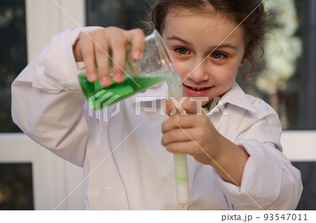Homeschooling Caucasian little girl in white lab coat learning about science, pouring reagent solution from flask into a test tube, watching fascination going on chemical reaction. Learning chemistry Homeschooling Caucasian little girl in white lab coat learning about science, pouring reagent solution from flask into a test tube, watching fascination going on chemical reaction. Learning chemistry 93547011
