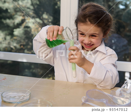 Cheerful little chemist, preschooler girl standing at the table and observing the chemical reaction going on in the glass flask with excited and surprised looks on her face. Miracles of chemistry Cheerful little chemist, preschooler girl standing at the table and observing the chemical reaction going on in the glass flask with excited and surprised looks on her face. Miracles of chemistry 93547012