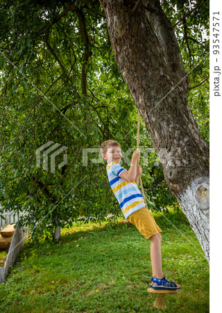 Happy little boy is having fun on a rope swing which he has found while having rest outside city. Active leisure time with children Happy little boy is having fun on a rope swing which he has found while having rest outside city. Active leisure time with children 93547571