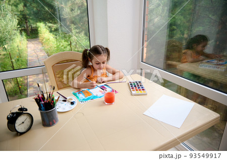 Adorable child, elementary age girl sitting at a table and painting picture with watercolors and paintbrush. Kids education and development. Drawing. Art classes and courses for telented children Adorable child, elementary age girl sitting at a table and painting picture with watercolors and paintbrush. Kids education and development. Drawing. Art classes and courses for telented children 93549917