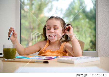 Cheerful Caucasian little girl of preschool, in orange top sitting at a table in the home veranda and drawing picture with paint brush and watercolors, enjoying art class for talented children Cheerful Caucasian little girl of preschool, in orange top sitting at a table in the home veranda and drawing picture with paint brush and watercolors, enjoying art class for talented children 93549918