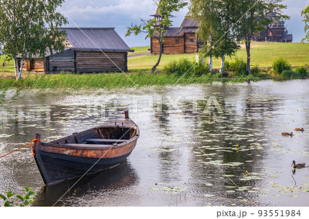 Landscape on Kizhi island in Lake Onega on a summer sunny day. Old wooden fishing boat Kizhanka on shore. Village with vintage mill, temple and church of Transfiguration in background. Karelia Russia 93551984