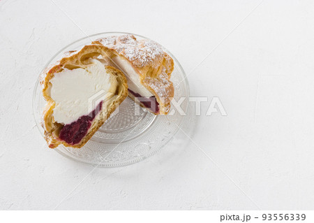 Choux Bun with whipped cream, cherry jam and sugar powder on top, cutted in half on glass plate, white background. Delicate choux pastry dessert. French cream puff. Close-up, copy space 93556339