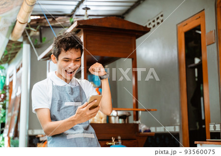 happy male merchant wearing apron looking at mobile phone screen 93560075