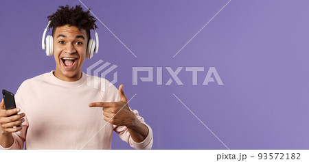 Technology and lifestyle concept. Portrait of happy, cheerful young man recommend awesome podcast or online music platform, buying subscribtion listen songs anytime, wear headphones, point at phone 93572182