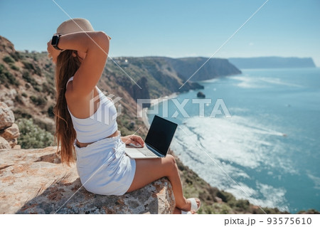 Digital nomad, woman in the hat, a business woman with a laptop sits on the rocks by the sea during sunset, makes a business transaction online from a distance. Freelance, remote work on vacation. Digital nomad, woman in the hat, a business woman with a laptop sits on the rocks by the sea during sunset, makes a business transaction online from a distance. Freelance, remote work on vacation. 93575610
