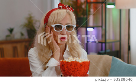 Woman sitting on couch eating popcorn and watching interesting TV serial, sport game online at home Woman sitting on couch eating popcorn and watching interesting TV serial, sport game online at home 93589640