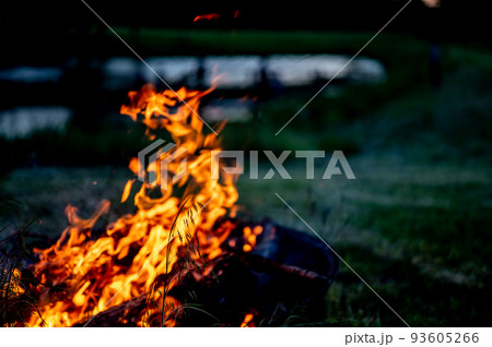 Leaping flames of a camp fire with a darkening view of a pond in the background. 93605266