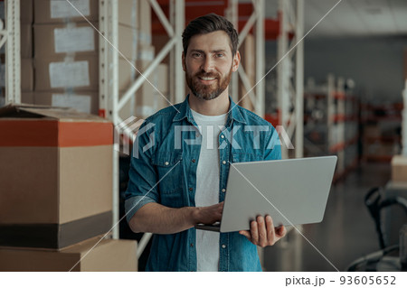 Handsome business owner working on laptop stands on own warehouse and looking at camera Handsome business owner working on laptop stands on own warehouse and looking at camera 93605652