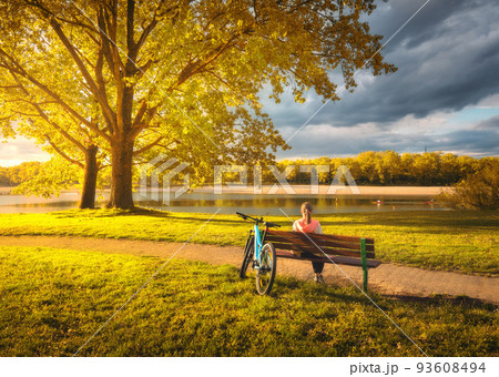 Woman sitting on bench and mountain bike, yellow trees and lake Woman sitting on bench and mountain bike, yellow trees and lake 93608494