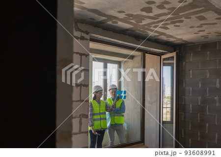 two engineers in protective vests and white helmets glue stickers on the window and discuss the construction plan of the facility or the scheme two engineers in protective vests and white helmets glue stickers on the window and discuss the construction plan of the facility or the scheme 93608991