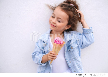 A girl in a denim blue jacket with a horn and a ball of pink ice cream straightens her hair flying in the wind, standing against a white concrete wall. Enjoying the milky creamy taste A girl in a denim blue jacket with a horn and a ball of pink ice cream straightens her hair flying in the wind, standing against a white concrete wall. Enjoying the milky creamy taste 93633398