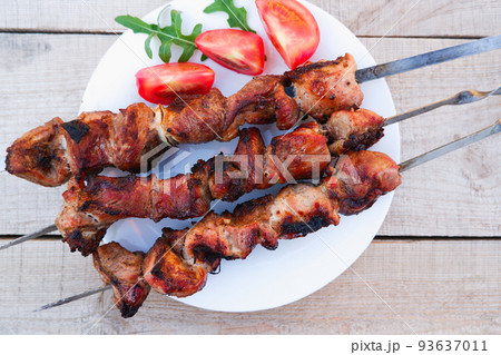 Juicy meat on skewers with tomatoes and herbs on a white plate on a wooden background. Kebab of meat is fried on the grill. Juicy meat on skewers with tomatoes and herbs on a white plate on a wooden background. Kebab of meat is fried on the grill. 93637011