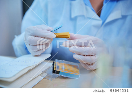 Details: A lab technician hands in medical gloves holding a container with litmus or reagent paper for measuring the PH, alkalinity and acidity of substances in chemical research laboratory. Close-up Details: A lab technician hands in medical gloves holding a container with litmus or reagent paper for measuring the PH, alkalinity and acidity of substances in chemical research laboratory. Close-up 93644551