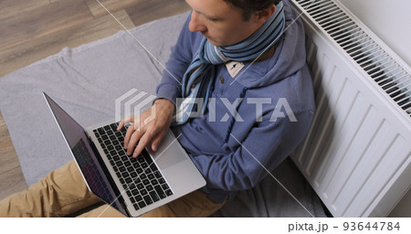 Man works on laptop near battery. Man freelancer in scarf warms himself near radiator. Sitting alone in room try warming indoor. Cold house, poor central heating, low temperature in apartment concept Man works on laptop near battery. Man freelancer in scarf warms himself near radiator. Sitting alone in room try warming indoor. Cold house, poor central heating, low temperature in apartment concept 93644784