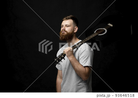 Young bearded guitarist in grey t-shirt holds a guitar on a shoulder in a dark room. 93646518