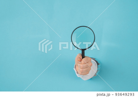 A woman's hand sticking out of a hole in a paper blue background holds a magnifying loupe. Copy space. A woman's hand sticking out of a hole in a paper blue background holds a magnifying loupe. Copy space. 93649293