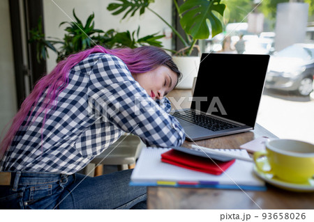 Young woman lying on her arms on the table in cafe in front of laptop with cup of coffee, sleepy. 93658026