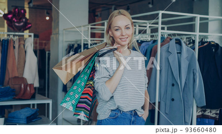 Portrait of attractive young woman standing with many colourful paper bags in modern clothing store, smiling happily and looking at camera. Stylish clothes is in background. 93660480