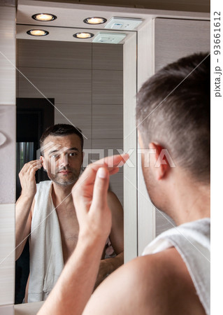 A man looks in the mirror in the morning and straightens his hair in the bathroom with a towel on his shoulder 93661612