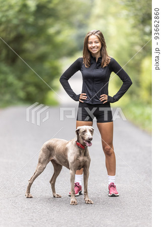Young sporting woman and her Weimaraner dog stand together in the forest Young sporting woman and her Weimaraner dog stand together in the forest 93662860