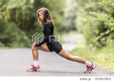 Young female athlete performs warm-up and legs stretching on a road in the nature Young female athlete performs warm-up and legs stretching on a road in the nature 93662863