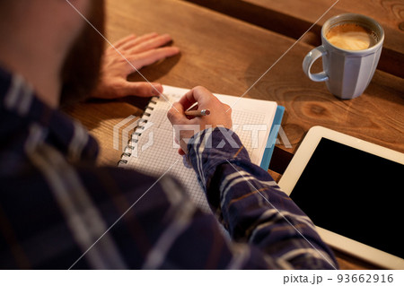 Young bearded businessman sits in cafe, home at table and writes in notebook, near lies tablet computer with black screen. Man is working, studying. 93662916