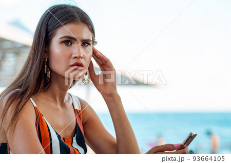 A woman with a tense expression is holding a phone in her hands. Summer cafe on the beach. Concept of business and social networks 93664105