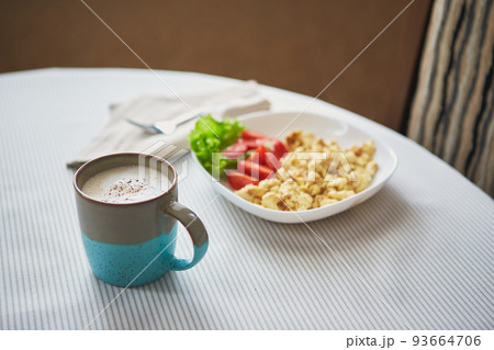 Cappuccino coffee in cup and plate with scrambled eggs and tomatoes. Breakfast on the table 93664706