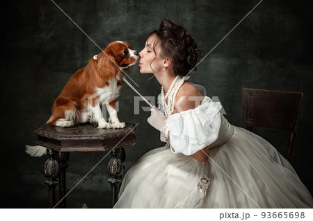 Kiss. Vintage portrait of young elegant woman in image of medieval person in renaissance style dress with little cute dog isolated on dark background. Kiss. Vintage portrait of young elegant woman in image of medieval person in renaissance style dress with little cute dog isolated on dark background. 93665698