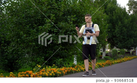 A young dad walks with a child sitting in a kangaroo backpack. A single father walks through the park with a phone in his hands. A young dad walks with a child sitting in a kangaroo backpack. A single father walks through the park with a phone in his hands. 93668518