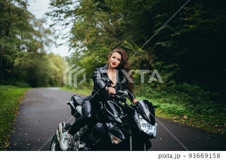 Portrait of a beautiful young woman sitting on a motorcycle, riding a motorcycle in black leather clothes on nature, street, forest, in sunny weather, in full growth 93669158