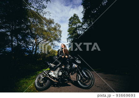 Portrait of a beautiful young woman sitting on a motorcycle, riding a motorcycle in black leather clothes on nature, street, forest, in sunny weather, in full growth Portrait of a beautiful young woman sitting on a motorcycle, riding a motorcycle in black leather clothes on nature, street, forest, in sunny weather, in full growth 93669693