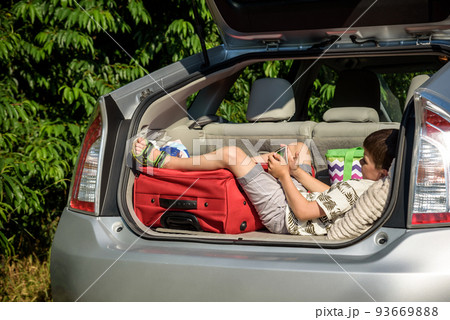 Cute little boy laying on the back of the bags and baggage in the car trunk ready to go on vacation with happy expression. Kid resting playing on smartphone 93669888
