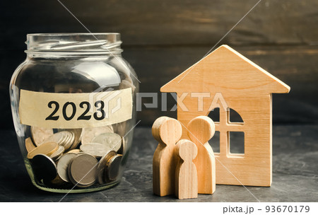 Family budget planning concept. Investments, plans, savings. Mortgage rates. Real estate. Glass jar with coins 2023 and family near the wooden house 93670179