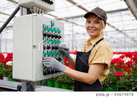 Pleasant woman in apron and cap presses the buttons on the  control panel of the automatic watering system for flowers in the greenhouse 93674078
