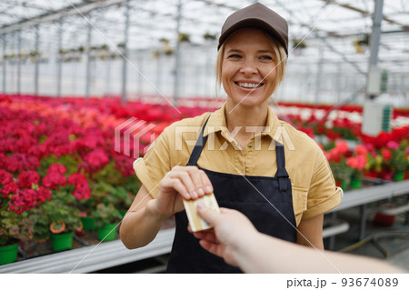 Portrait of a friendly woman seller a greenhouse with flowers taking a payment card from a customer Portrait of a friendly woman seller a greenhouse with flowers taking a payment card from a customer 93674089