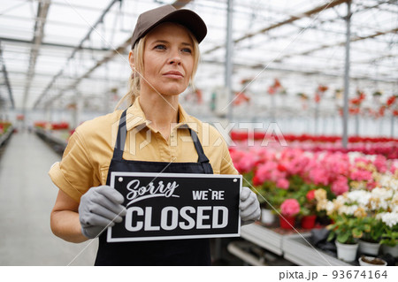 Pretty middle aged woman in gardener uniform holding sign we are closed in greenhouse 93674164
